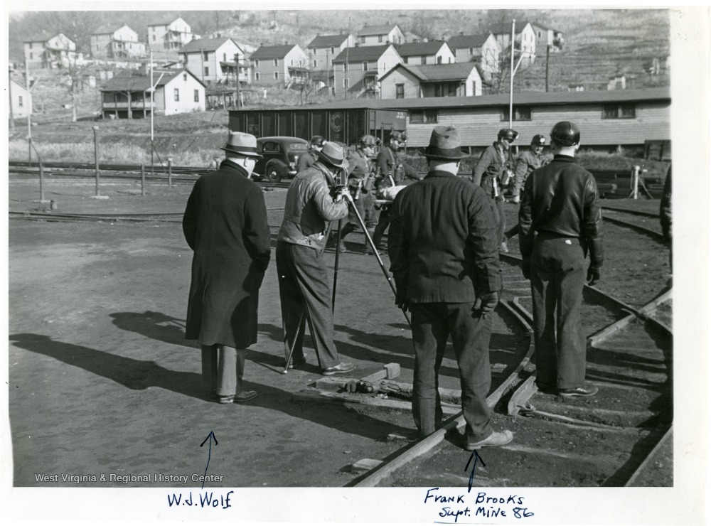 Miners carrying an injured person on a stretcher while a man films. W.J. Wolf and Frank Brooks the Supt. of Mine 86 are included in the picture.