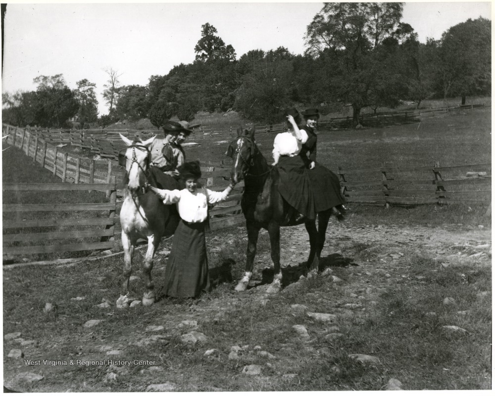 Four women on horses, two on each horse and a woman standing in between them.