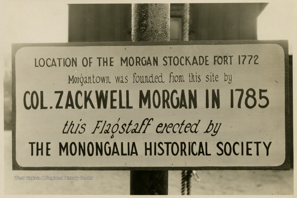 The sign reads 'Location of the Morgan Stockade Fort 1772 Morgantown was founded from this site by Col. Zackwell Morgan in 1785 this Flagstaff erected by The Monongalia Historical Society'.