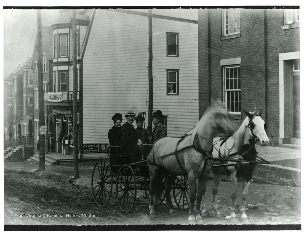 Two horses are pulling a wagon with three ladies and a gentleman down Pleasant Street in Morgantown, West Virginia.