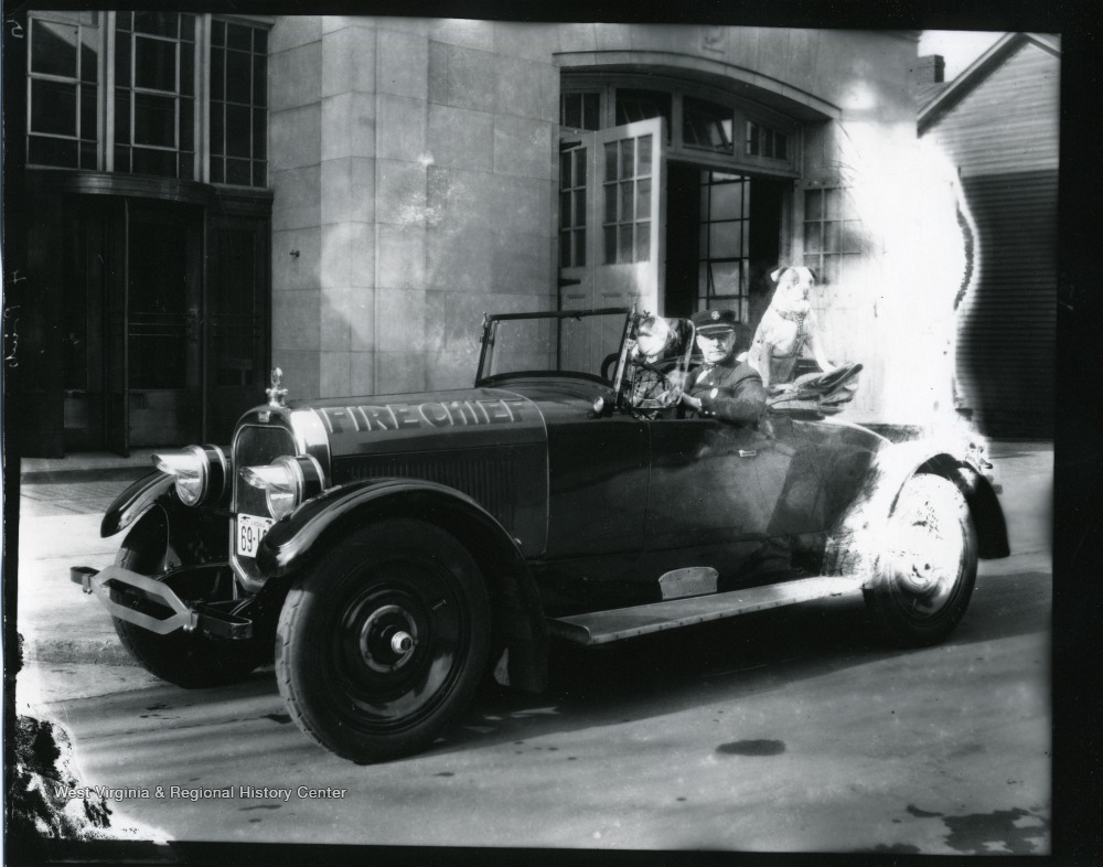 The car, which reads "Fire Chief," is parked in front of the Morgantown Fire Department building on Spruce Street.  Doc, the fire department mascot, is in the backseat of the car.
