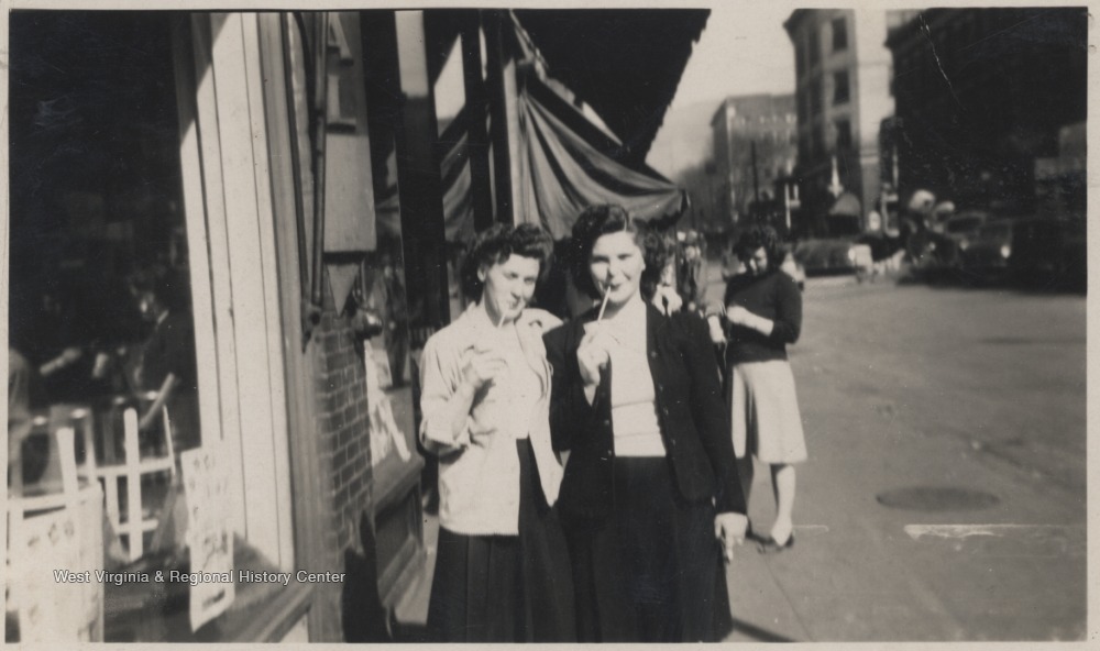 Tommy Smith Trail and Edith Jeffries Adkins stand outside a building drinking from straws. 