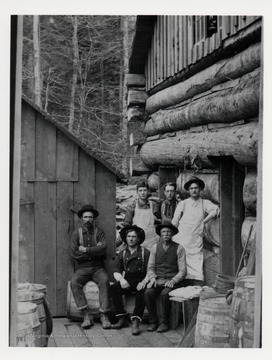 Six men pose beside a camp building.  
