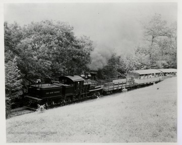 Cass Scenic Railroad.  Train engine with loaded passenger cars on tracks beside hill and forest.  
