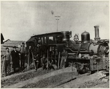 Crew members pose beside train engine pulling lumber car.