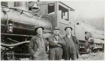 Three men standing in front of a train engine pulling lumber cars.