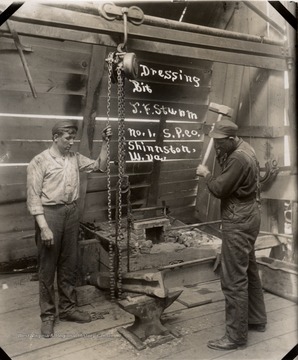 Two men forging a drill bit, one holding chain, one raising hammer.