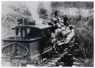 Climax train engine No. 2 pulling log carts.  Crew members standing on top of the logs and one sitting on top of the engine.