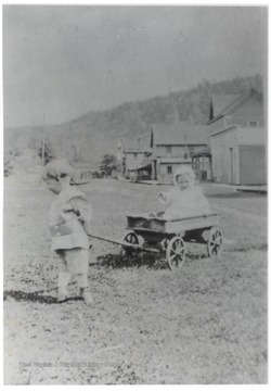 A small child pulling a wagon through the grass with another small child in it.