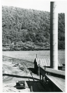 A towering smokestack to the right with railroad tracks running down the center and in the distance.    A truck sits in the center with its door open.