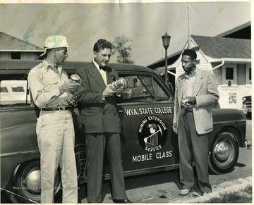 Three men inspecting lanterns next to a W. Va. State College Mining Extension Service Mobile Class car. 