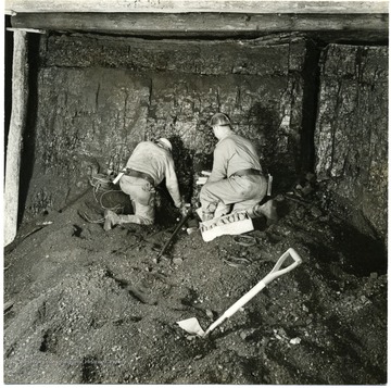 Two men placing charges in an already drilled hole. Notice the preparatory cut on upper right hand side. 