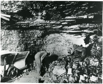 Two miners work on a seam, one with a pick, the other with a shovel.