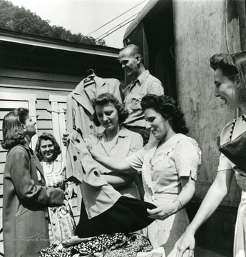 Ladies examine dresses and coats for sale.  Man holding coat at center top of picture is D.M. Hughes who was the driver of the truck and sold clothes.