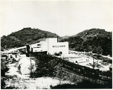 'An overall view of the new plant at Williams No. 98 Mine of Consolidation Coal Compnay (W. Va.) is shown. Coal comes from the mine on a 558 foot conveyor belt at the left to the top of the plant past the old tipple (shown dark against the new structure) which is now being removed. Through intricate processes it is screened and sized and loaded into railroad cars on four tracks which pass under the tipple (center). Impurities removed from the coal are carried on another long conveyor to a hilltop slate dump shown in the extreme upper right corner. This mine is named for Eugene Williams of Romney, W. Va. Mr. Williams is Chairman of the Board of Western Maryland Railroad. Williams Preparation Plant, convering over three acres of floor space on eight levels is a steel and concrete structure 100 feet high and fully enclosed without a single window. this windowless 'factory' is a mystifying innovation to all who visit the plant. It is equipped with a ventilating system, fluorescent lighting, and a complete vacuum type dust collection system that gather in all air borne particles at their source. All of these measures promote health, safety and good housekeeping. The raw coal is delivered to the preparation plant at a rate of 450 tons per hour by a 558 foot belt which picks up the coal at the rotary mine car dump at the bottom of the slope. After the coal is crushed down to 5 inch top size the raw coal is fed into a 15 foot diameter chance cone yielding 278 tons of 'float-kleen' coal per hour. The washing process operates on the principle of gravity separation, a fluid mass of sand and water being utilized as the density medium. Designed and built by Fairmont Machinery Company placed in operation March 31, 1953;  No windows, dust collection system; track storage, 100 empties, 80 loads; Present daily capacity, 8500 tons; potential daily capacity, 15,000 tons; coal reserves, 80,000,000 tones, Pittsburgh seam; slack dried, centrifugal and thermal dryers; oil treating facilities for stoker grades; served by two railroads, WM and B&amp;O; Plant named for Eugene Williams of Romney, W. Va. Board Chairman of the Western Maryland Railroad.'