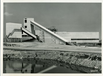 'The Georgetown Prepatation Plant is here viewed from the north, showing the main entrance. Landscaping has begun and the company will build a sizable parking lot in front of the plant with auxiliary lots near the scale house (off the picture to the left) and at the track level in the rear. The enclosed raw coal conveyor and the refuse loading bin are shown at the right. This plant will serve as a central preparation facility for the strip mining and some of the underground mining operations of the Hanna Coal Company, Ohio operating division of Pittsburgh Coal Company. With a capacity of 1,500 tons of raw coal input per hour, Georgetown is the largest coal cleaning plant in the commerical bituminous industry. It has three cleaning circuits and five types of facilities for drying the coal. Integrated with the plant operation are special systems for cleaning and recirculating the water, thereby avoiding stream pollution, and for elimination of air pollution.'