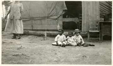 Three African American children sitting on the ground near a home and a woman on the left.