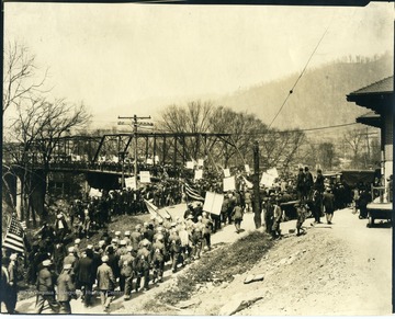 Many miners parading down the road and accross a steel bridge while carrying signs and American flags.