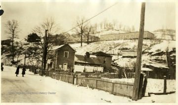 View of barracks on hill above house with picket fence. Two females are standing in what appears to be the snowy road. 