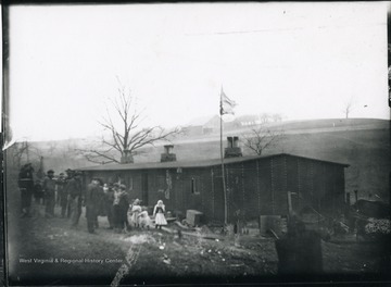 Group of people stand outside of the barracks. Barn in a field in the distance.