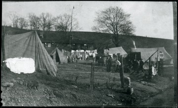 Tents behind fence in front of building. Laundry hanging on line. Some men on right side.