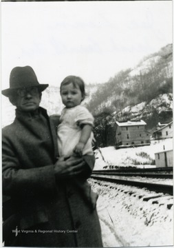 Man holding little girl with train tracks, houses, and snow covered hillside in the background.