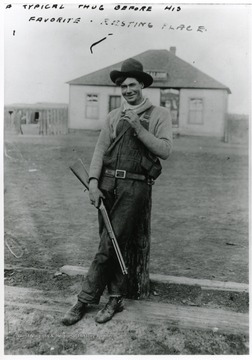A man standing with his gun leaning against a post, Saloon in the background.