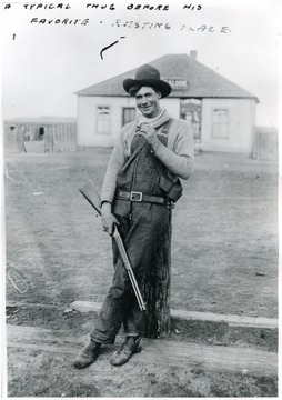 A man with a gun leaning against a post, saloon in background.