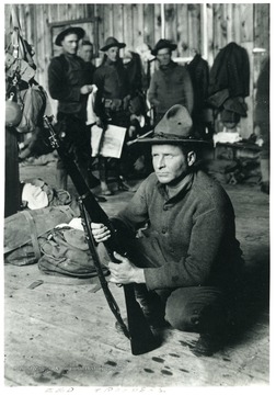 A man kneeling with his rifle.  Other men standing in the background. 