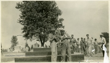 Two men smile for the camera in front of the foundation for the Mother Jones monument.  A child stands on the monument and a group of men are gathered behind it.