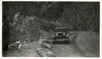 Car parked along road beside a hillside.  Man standing on the side of the picture.