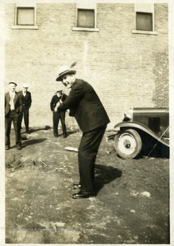 Man wearing a hat getting ready to swing a bat-like item.  Three men in the background watching. 