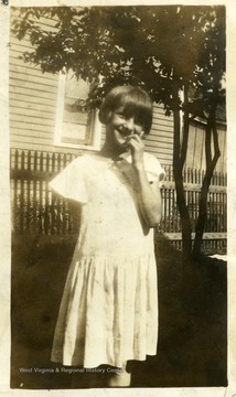 Young girl standing in front of a house smiling with her hand near her mouth. 