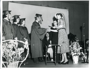 Mrs. Eleanor Roosevelt shakes the hand of a graduate and hands out diplomas during the graduation ceremony at Arthurdale High School in Arthurdale, West Virginia.  Jennings Randolph visible on the stage.