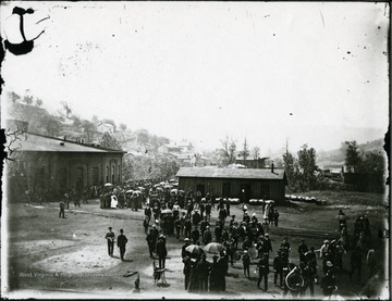 Crowds of people in between a brick and wooden building. A band is visible in the bottom right corner of picture.