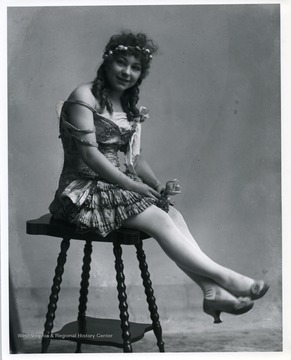 Woman wearing a dress and holding a flower is sitting on a stool.
