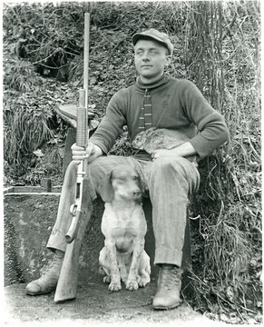Paul Aegerter sitting with a rabbit on his lap and a gun in his hand.  A dog is sitting beside him.  Helvetia, W. Va.