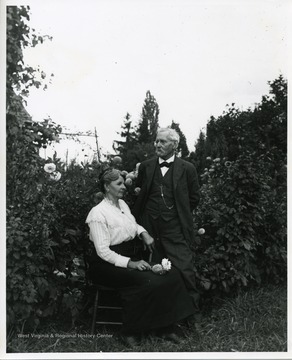 Elka Hassig is sitting near a flower garden while Ernest Hassig stands next to her.