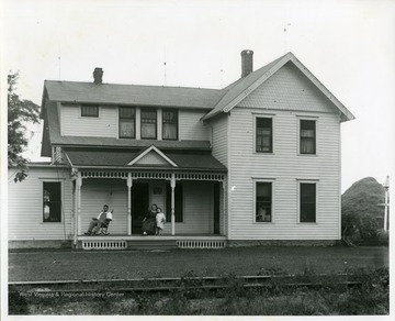 A man is sitting in a rocking chair while a lady is holding a young child on the front porch of a two-story house thought to be in West Virginia.
