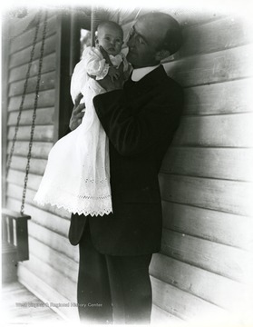 A man, possibly Benjamin Holtkamp, in a suit holds a baby in a long white dress.
