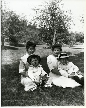 Two mothers holding small children while sitting in a field.