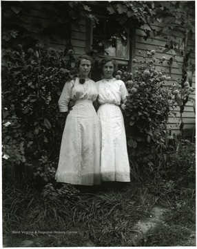 Two women standing together between blossoming dahlias, Helvetia, W. Va.