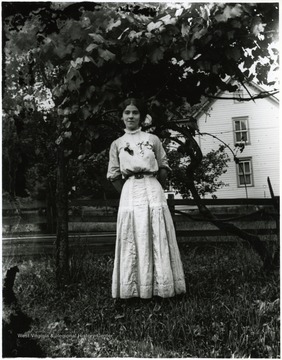 Young woman standing alone in front of a tree.  House in the background.  Helvetia, W. Va.