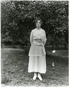 Young woman with a flag pinned to her dress standing alone in front of a tree in a fenced area.  Helvetia, W. Va.