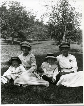 Two mothers are sitting with their children in a field near some trees.