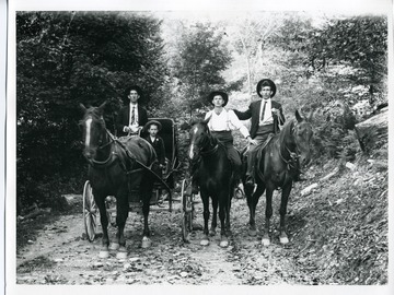 Two men are riding horses while another man and a young boy are riding in a horse drawn carriage down a dirt path.