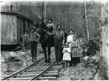 Man is holding horse's bridle in the center of picture with little boy riding the horse.  Lumber camp bunkhouses are visible at left.
