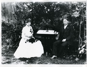 Clara Koerner and John Gobeli are sitting at a table. Clara is wearing a long white dress and is holding flowers while John is wearing a three piece suit. 