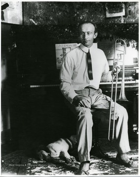 Man holding a trombone sitting beside a piano while a dog is sleeping beside him.