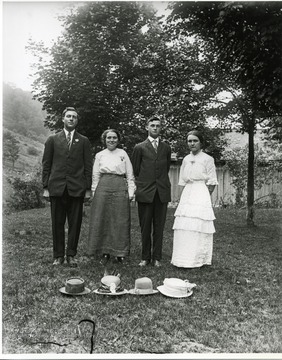 Two gentlemen wearing three piece suits are joined with two ladies wearing long dresses. Four hats are on the ground in front of them.